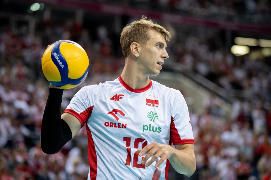 Siatkarz w biało-czerwonym stroju polskiej reprezentacji przygotowuje się do zagrywki podczas meczu, w tle widoczna publiczność na trybunach.