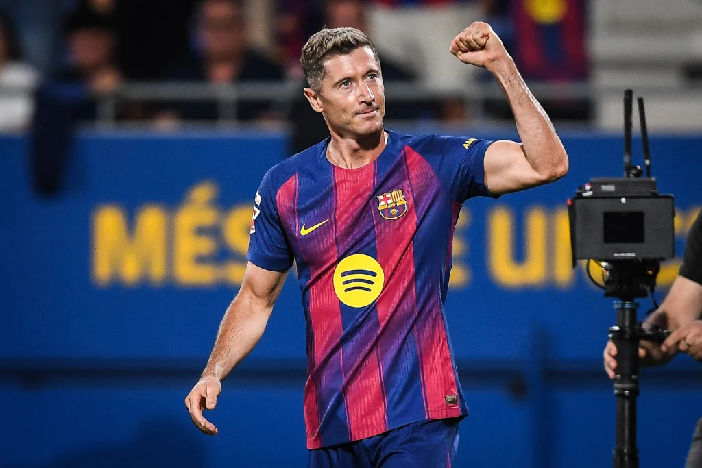 Piłkarz w stroju FC Barcelony unosi zaciśniętą pięść w geście triumfu, na tle stadionu i kamery transmisyjnej, z wyraźnie widocznym logo sponsora na koszulce.