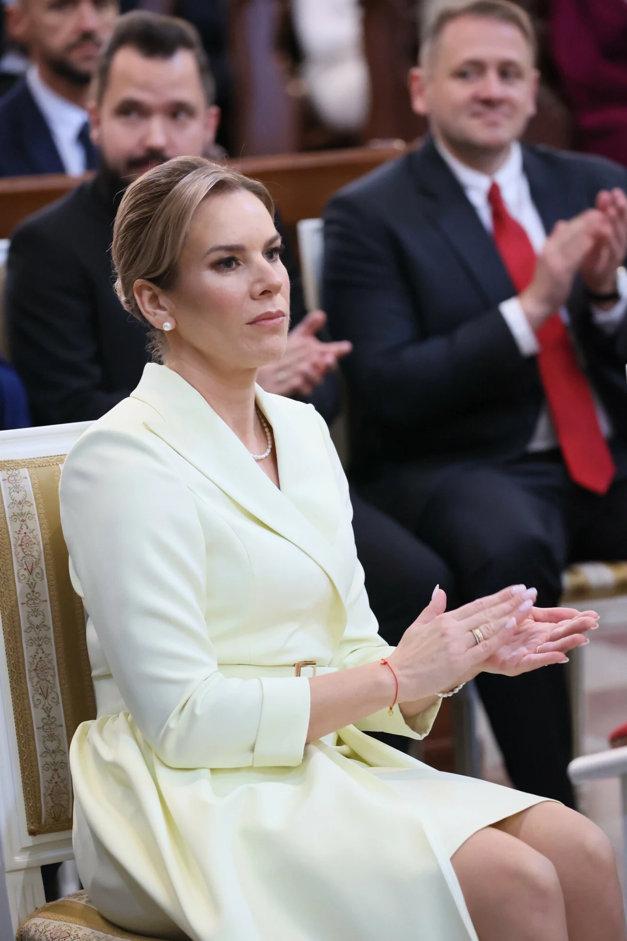 Elegancko ubrana kobieta siedząca na krześle, klaszcząca w dłonie podczas oficjalnego wydarzenia, w tle widoczni inni uczestnicy w eleganckich strojach.