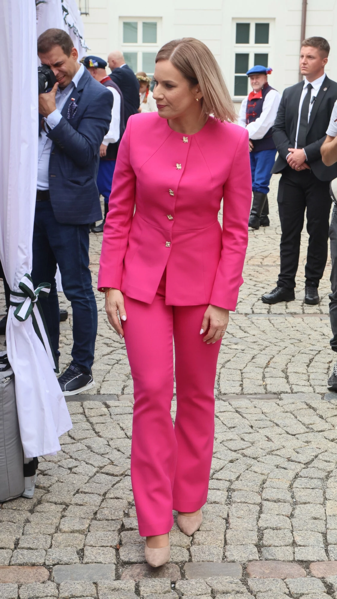 Dożynki Prezydenckie. Marta Nawrocka w różowym garniturze Kobieta ubrana w elegancki, różowy garnitur stoi na brukowanym placu, wokół niej widoczne są osoby w garniturach i mundurach, w tle biała elewacja budynku oraz zgromadzeni uczestnicy wydarzenia.