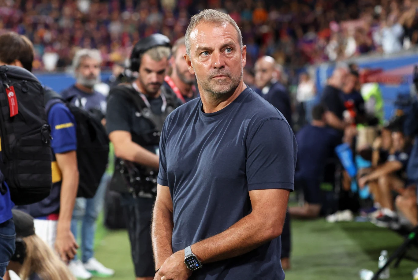 Hansi Flick Mężczyzna w granatowej koszulce stoi na murawie stadionu otoczony przez pracowników i reporterów podczas wydarzenia sportowego, na drugim planie widać tłum widzów na trybunach.