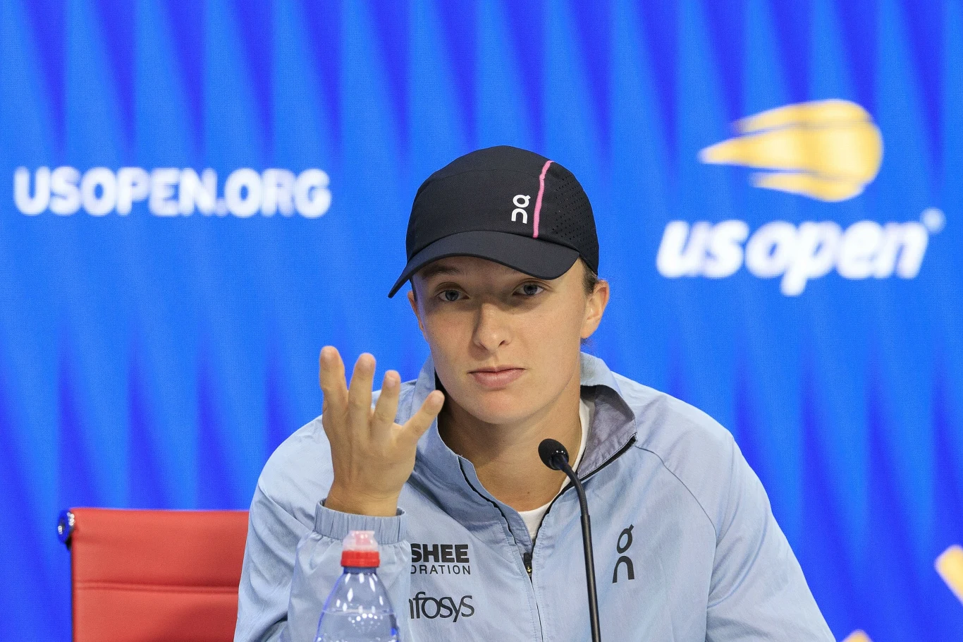 Iga Świątek Tenisistka podczas konferencji prasowej, ubrana w sportową bluzę i czapkę z daszkiem, siedzi przy mikrofonie na tle niebieskiego baneru US Open.