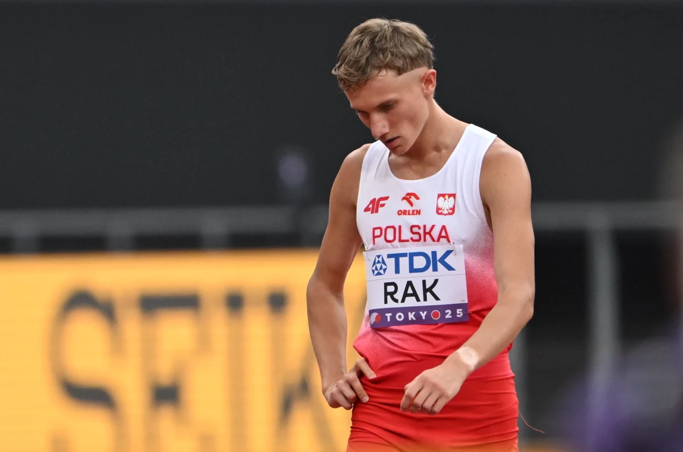 Lekkoatleta w stroju reprezentacji Polski na tle rozmytego stadionu, skupiony i patrzący w dół.
