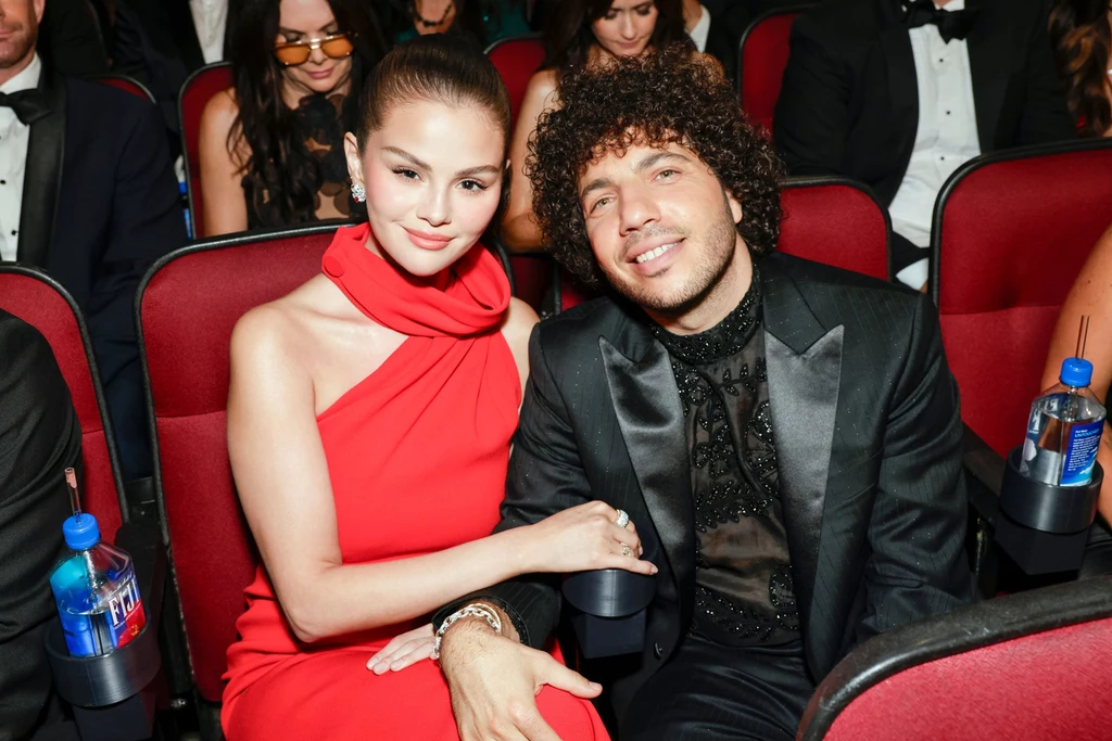 Selena Gomez promieniała u boku ukochanego Para ubrana elegancko, kobieta w czerwonej sukni i mężczyzna w czarnym garniturze, siedzą razem w rzędzie sali podczas wydarzenia, trzymając się za ręce.