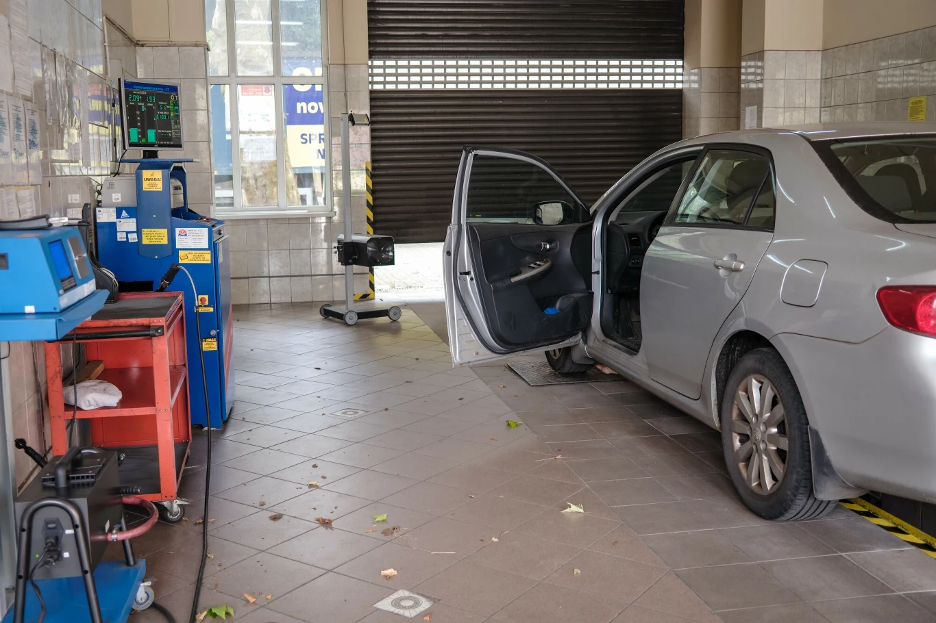 19 września znacznie wzrosną ceny badań technicznych. Posiadacze aut z LPG zapłacą 245 zł Szary samochód osobowy zaparkowany w warsztacie samochodowym z otwartymi drzwiami kierowcy, obok stanowisko diagnostyczne z komputerami i sprzętem warsztatowym, w tle widoczny zamknięty segmentowy garaż