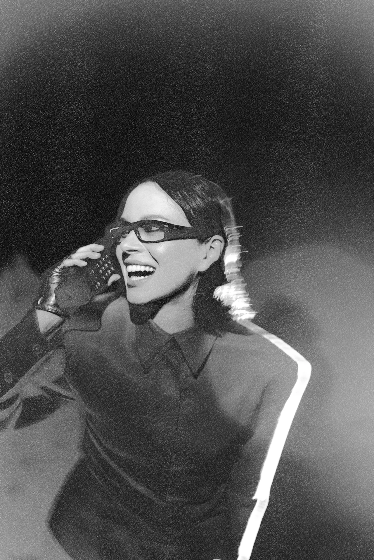 Kasia Lins jako Obywatelka K.L. Uśmiechnięta kobieta w okularach i ciemnej koszuli rozmawiająca przez telefon, stojąca na tle dymu lub mgły, w dynamicznym oświetleniu i z efektem rozmycia.