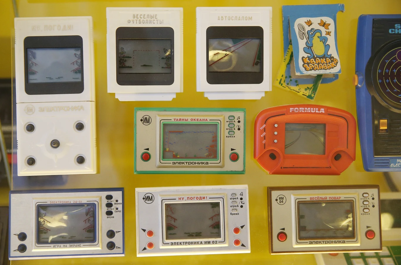 Kolekcja radzieckich gier elektronicznych z lat 80. i 90., wszystkie ułożone równo na żółtym tle; gry o różnych kształtach i kolorach z widocznymi ekranami LCD oraz rysunkami lub napisami na obudowach.