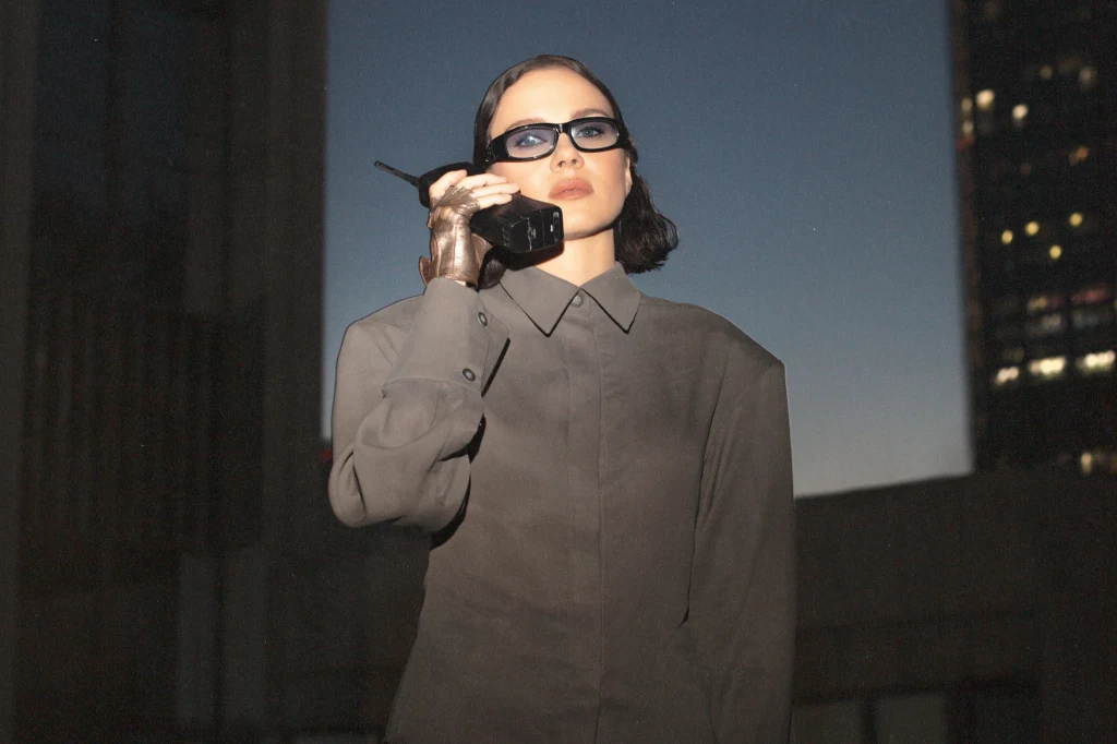 Kasia Lins w najnowszym projekcie, jako Obywatelka K.L. Kobieta ubrana w szarą koszulę z długim rękawem, nosząca ciemne okulary, trzyma staromodny telefon komórkowy przy uchu, stoi na tle wieżowca o zmierzchu.