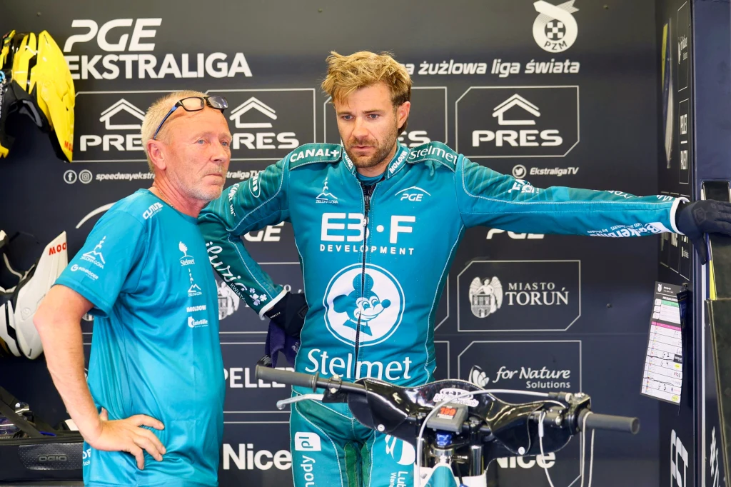 Dwóch mężczyzn ubranych w stroje żużlowe w strefie zawodników, jeden z nich opiera się o motocykl, w tle reklamy sponsorów i logotypy ligi żużlowej.