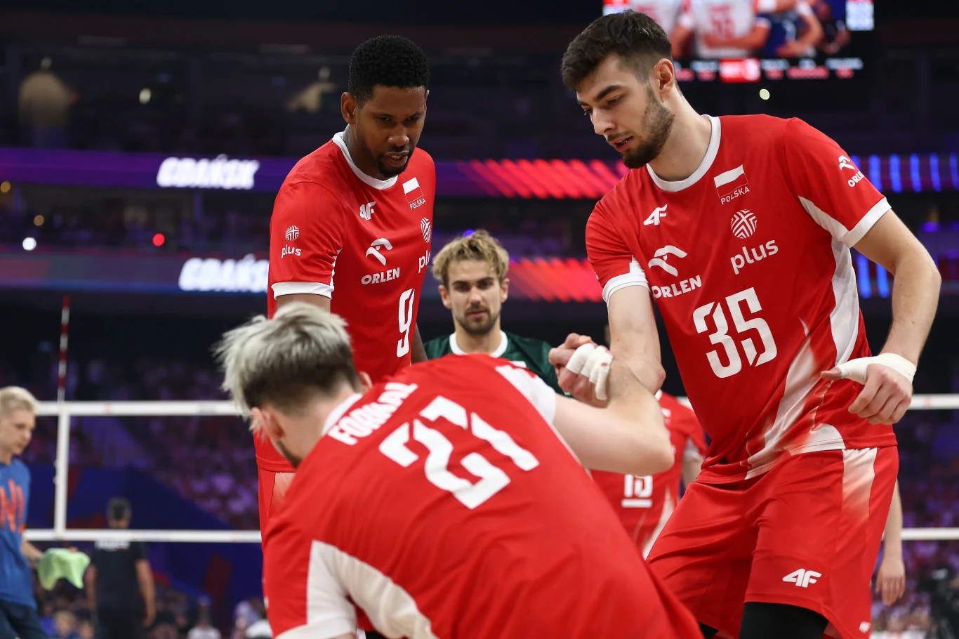 Zawodnicy siatkarskiej reprezentacji Polski w trakcie meczu, jeden z nich pomaga podnieść się koledze z drużyny na boisku, wszyscy ubrani w charakterystyczne czerwone stroje z białymi napisami i numerami.