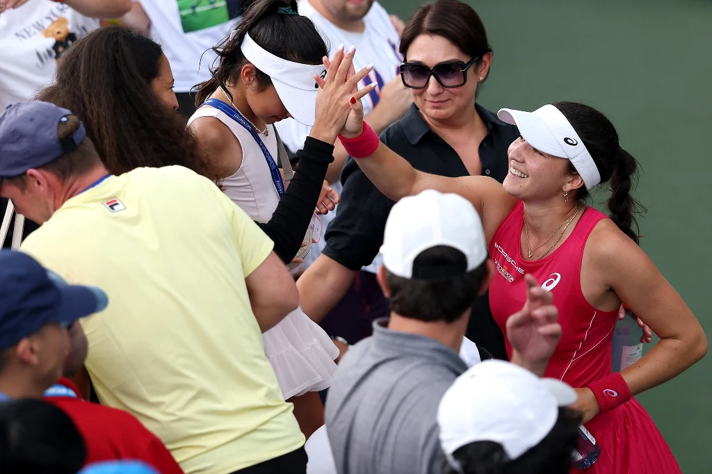 Eva Lys Tenisistka w czerwonym stroju przybija piątkę z fanami i osobami towarzyszącymi przy korcie, uśmiechnięta, w otoczeniu ludzi w sportowych strojach, panuje radosna atmosfera.