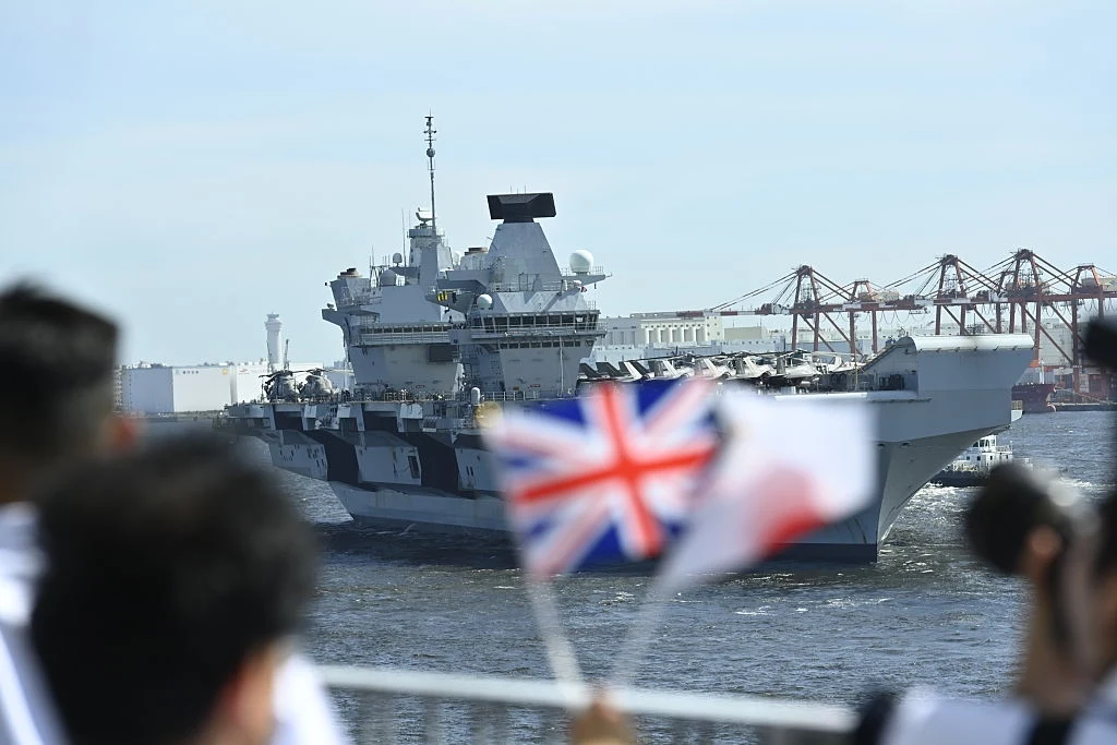Korea Południowa, USA i Japonia zacieśniają współpracę Lotniskowiec wojskowy wpłynął do portu, na pierwszym planie kibice machają brytyjską flagą oraz biało-czerwoną flagą, w tle widoczne dźwigi portowe.