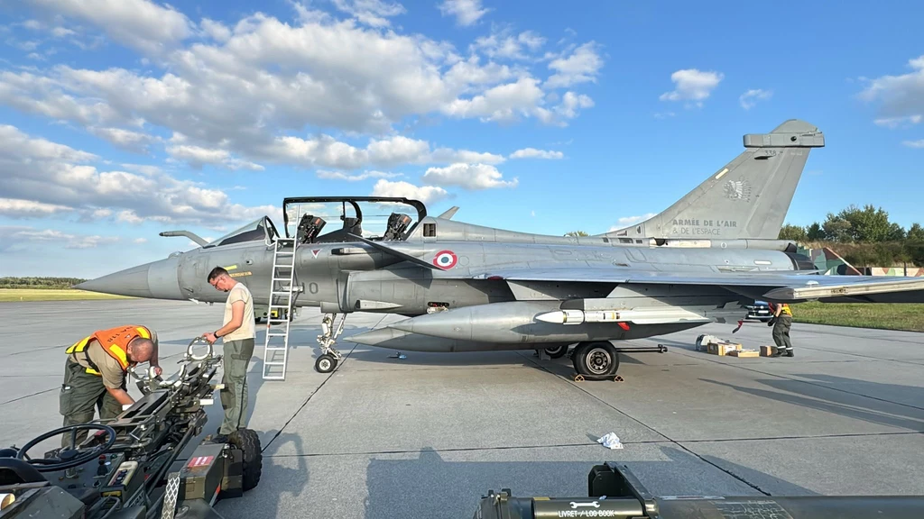 Myśliwiec wojskowy Rafale stoi na pasie lotniska, obok znajduje się dwóch techników prowadzących konserwację, po lewej stronie aparat do obsługi naziemnej, niebo częściowo zachmurzone.