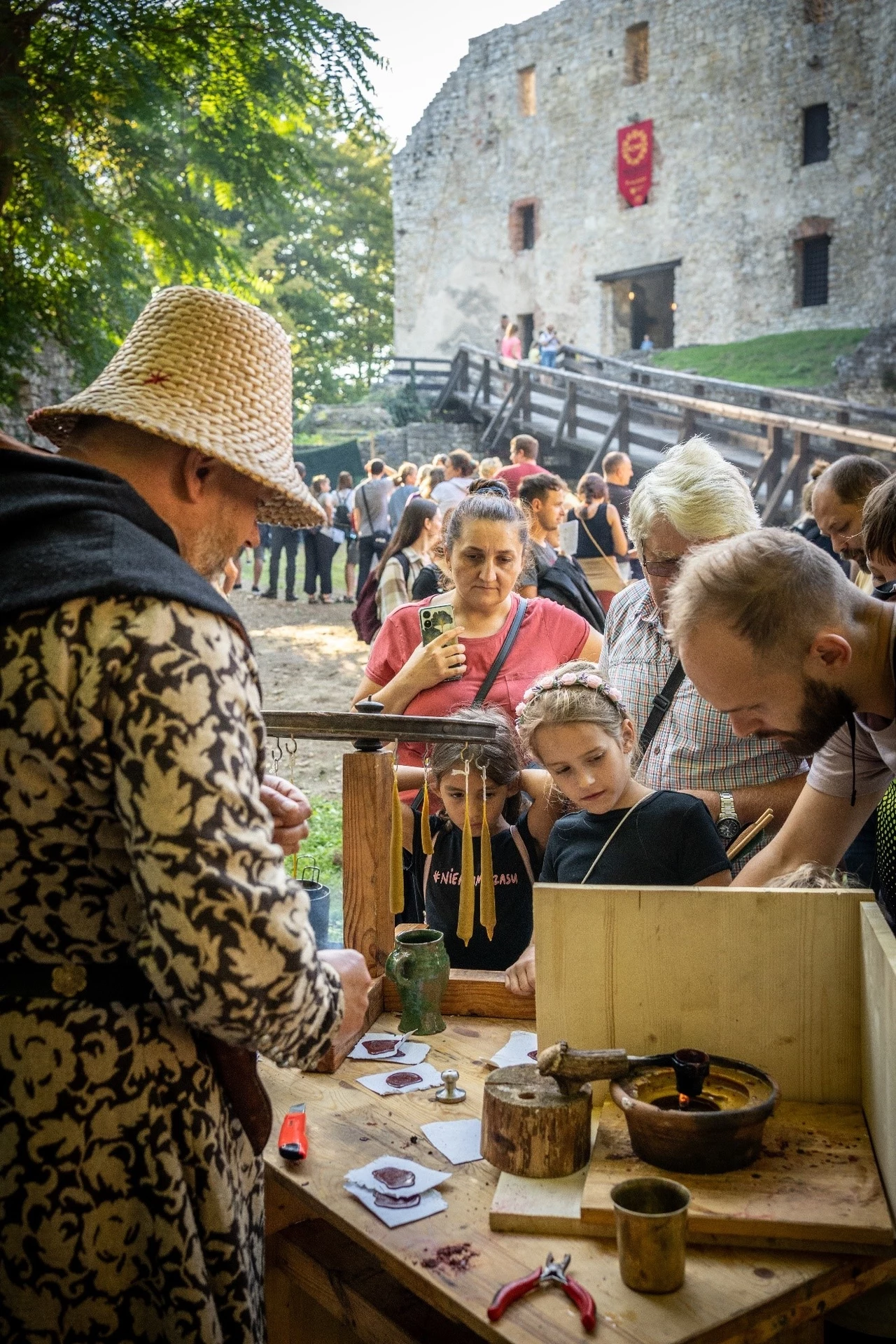 Grupa ludzi uczestniczących w warsztatach rzemieślniczych na dziedzińcu zamku, mężczyzna w stroju historycznym prowadzi pokaz, dzieci i dorośli uważnie obserwują, w tle widoczna kamienna budowla i zgromadzeni widzowie.