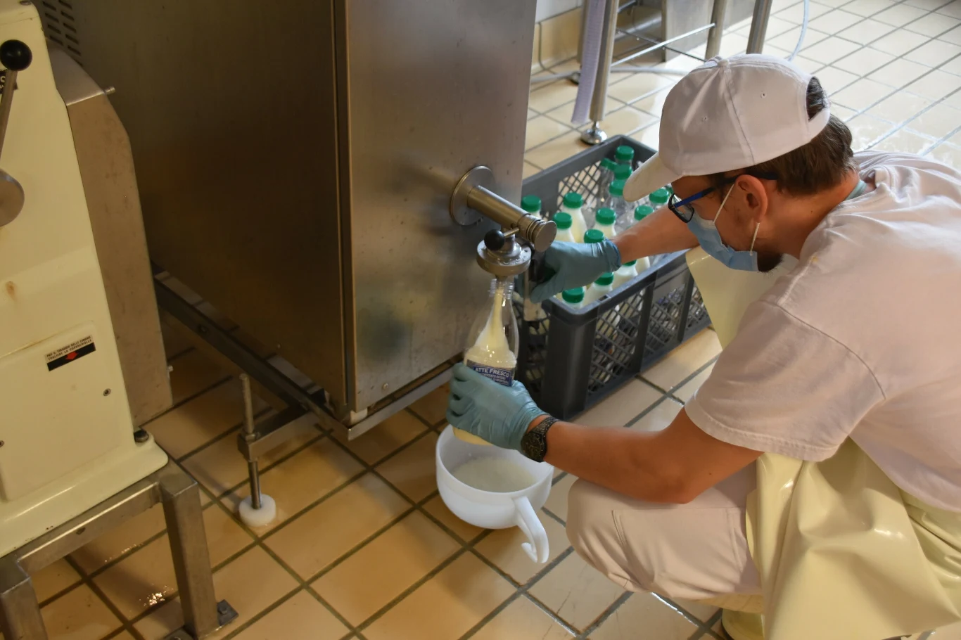 Polmlek wygasza produkcję w Szczekocinach. Wiadomo, co z pracownikami (zdj. ilustracyjne) Pracownik w sterylnym stroju pobiera mleko do butelki z dużej maszyny, obok pojemnik z pełnymi butelkami mleka, pomieszczenie wykafelkowane.