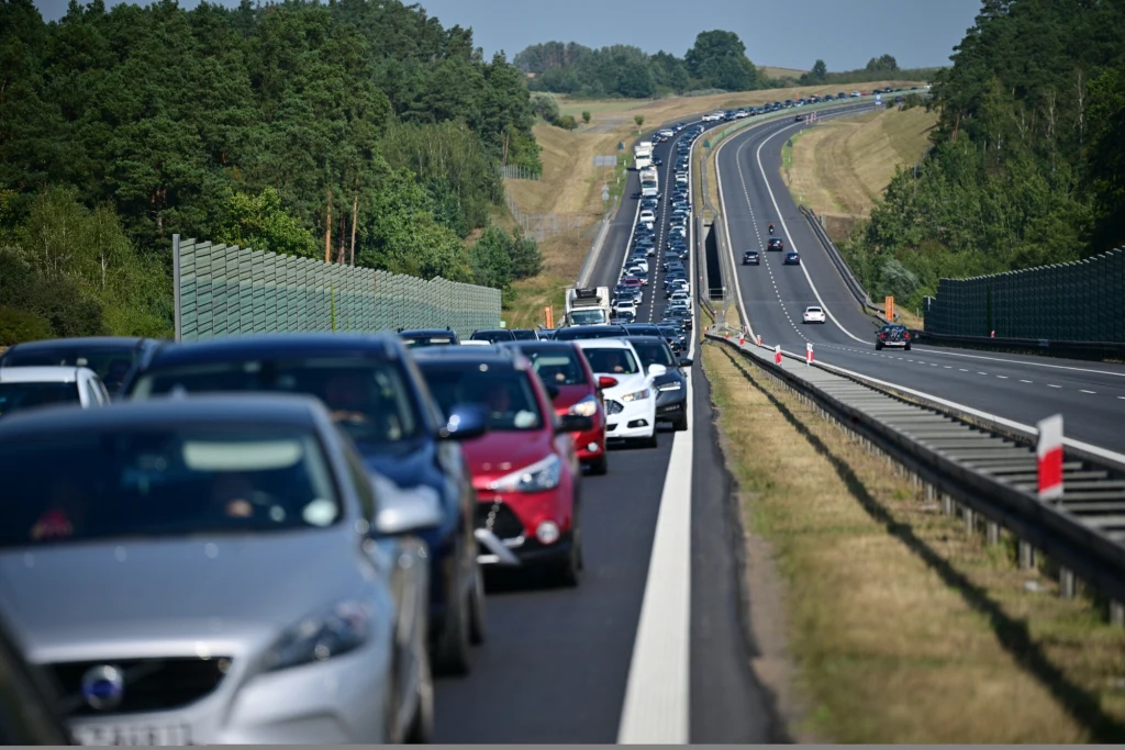 Długi korek samochodów na autostradzie otoczonej lasem, pojazdy stoją w obu kierunkach, a droga biegnie przez pagórkowaty, zielony krajobraz