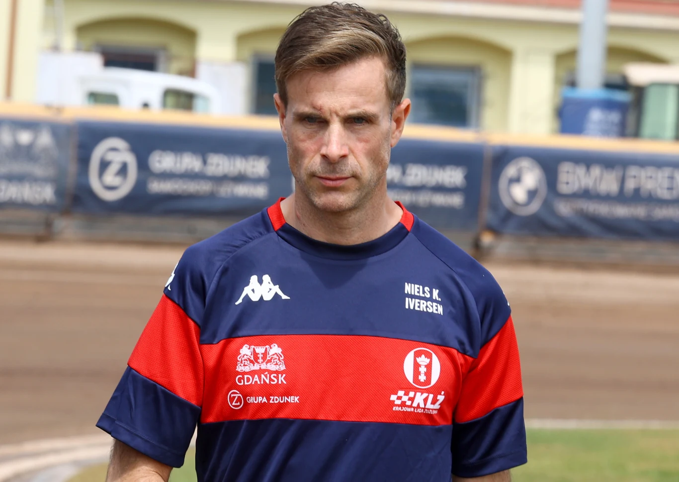 Mężczyzna w sportowej koszulce z napisem Niels K. Iversen, idzie po torze żużlowym, w tle widać bandy reklamowe oraz fragmenty budynków.