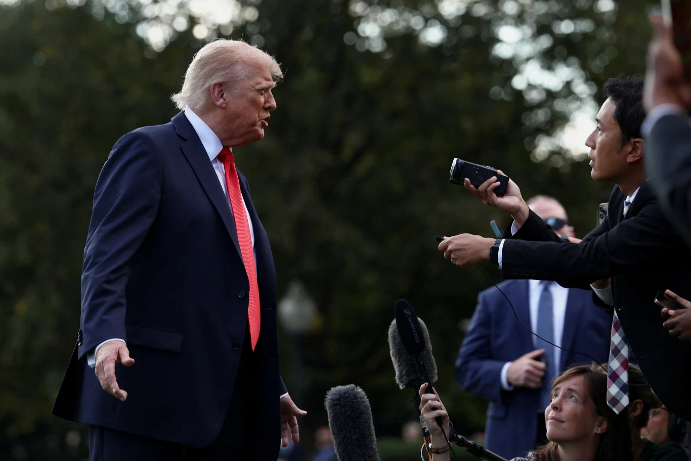 Prezydent USA Donald Trump Mężczyzna w garniturze udziela wywiadu dziennikarzom, którzy trzymają mikrofony i dyktafony w jego kierunku, w tle rozmyte zieleń drzew.