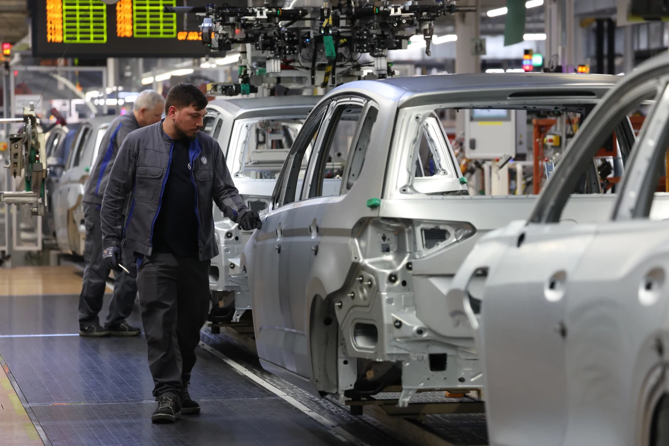 Premiera elektrycznego Volkswagena opóźniona | Fot. ilustracyjna Pracownicy fabryki samochodów kontrolują montaż karoserii pojazdów na linii produkcyjnej, jedna osoba uważnie przygląda się detalom surowego nadwozia, wokół widać maszyny i inne stanowiska robocze.