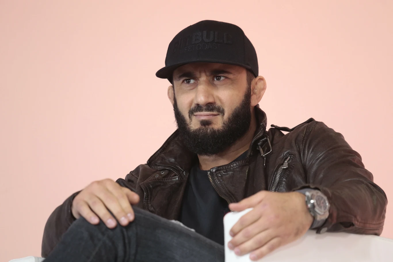 Mężczyzna z brodą ubrany w skórzaną kurtkę i czarną czapkę z daszkiem, siedzi na białym tle, patrząc przed siebie z lekko zmarszczonymi brwiami; na lewym nadgarstku nosi sportowy zegarek.