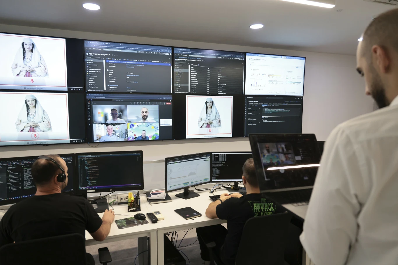 Zespół mężczyzn pracujący w centrum monitoringu przy komputerach, nad nimi kilka dużych ekranów wyświetlających rozmaite dane, obrazy osób i wykresy.