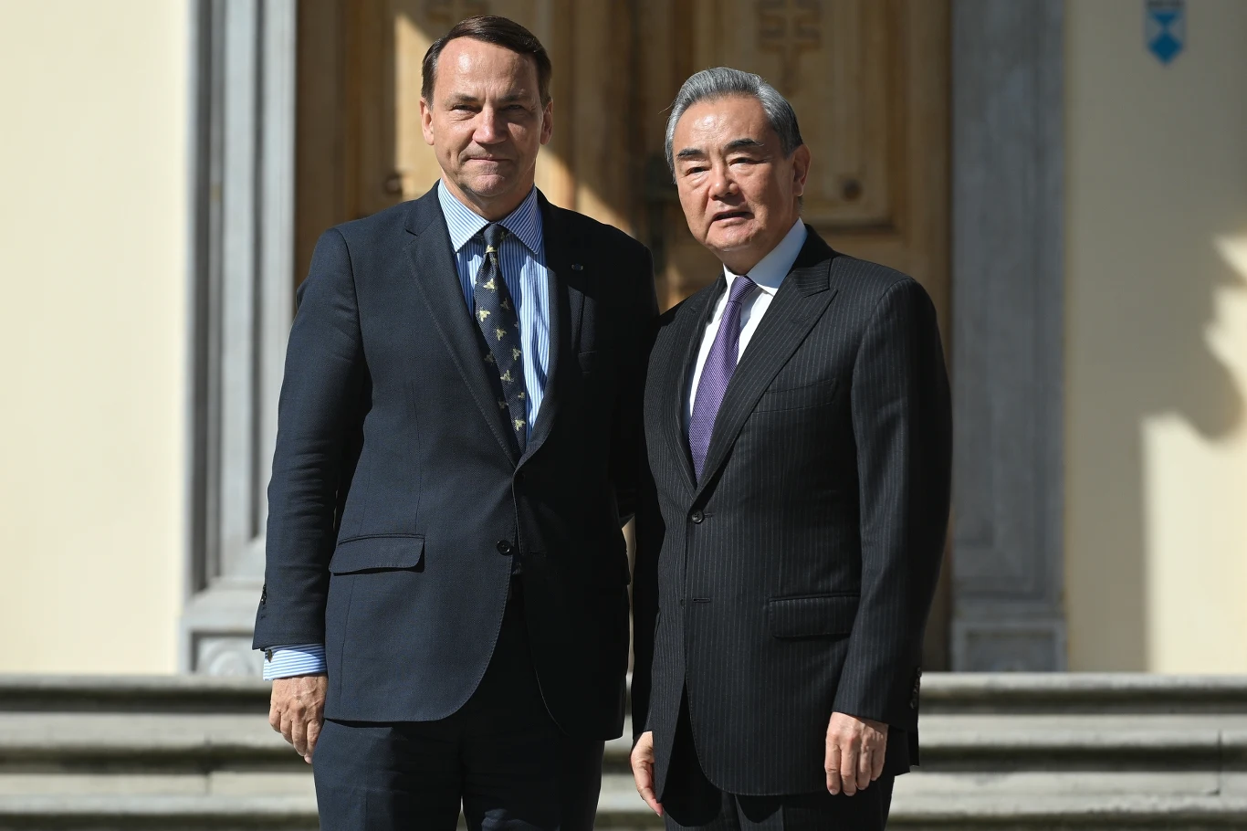 Dwóch mężczyzn w garniturach stoi przed wejściem do okazałego budynku, pozują razem na tle kamiennych schodów i zdobionych drzwi.