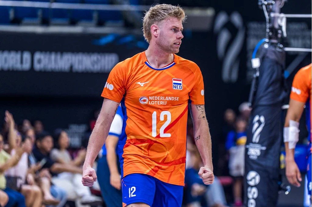Bennie Tuinstra, przyjmujący reprezentacji Holandii Siatkarz w pomarańczowym stroju z numerem 12, grający w reprezentacji Holandii podczas meczu siatkówki, z wyrazem determinacji na twarzy, w tle widownia i fragment siatki.