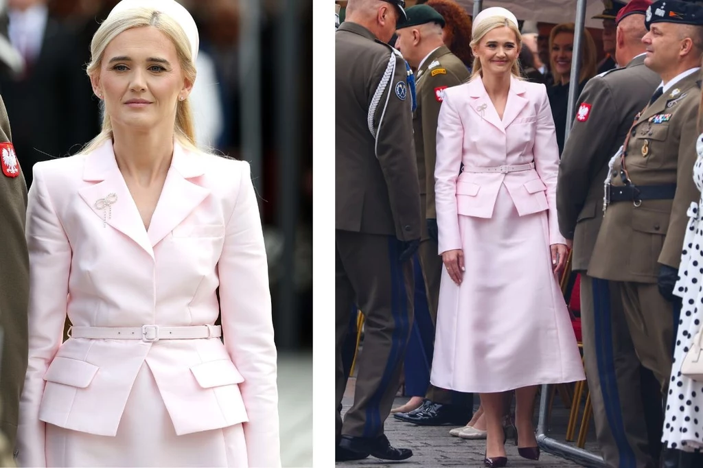 Kobieta w eleganckim, jasnoróżowym kostiumie, stojąca wśród żołnierzy w mundurach oraz innych osób uczestniczących w oficjalnym wydarzeniu na świeżym powietrzu.