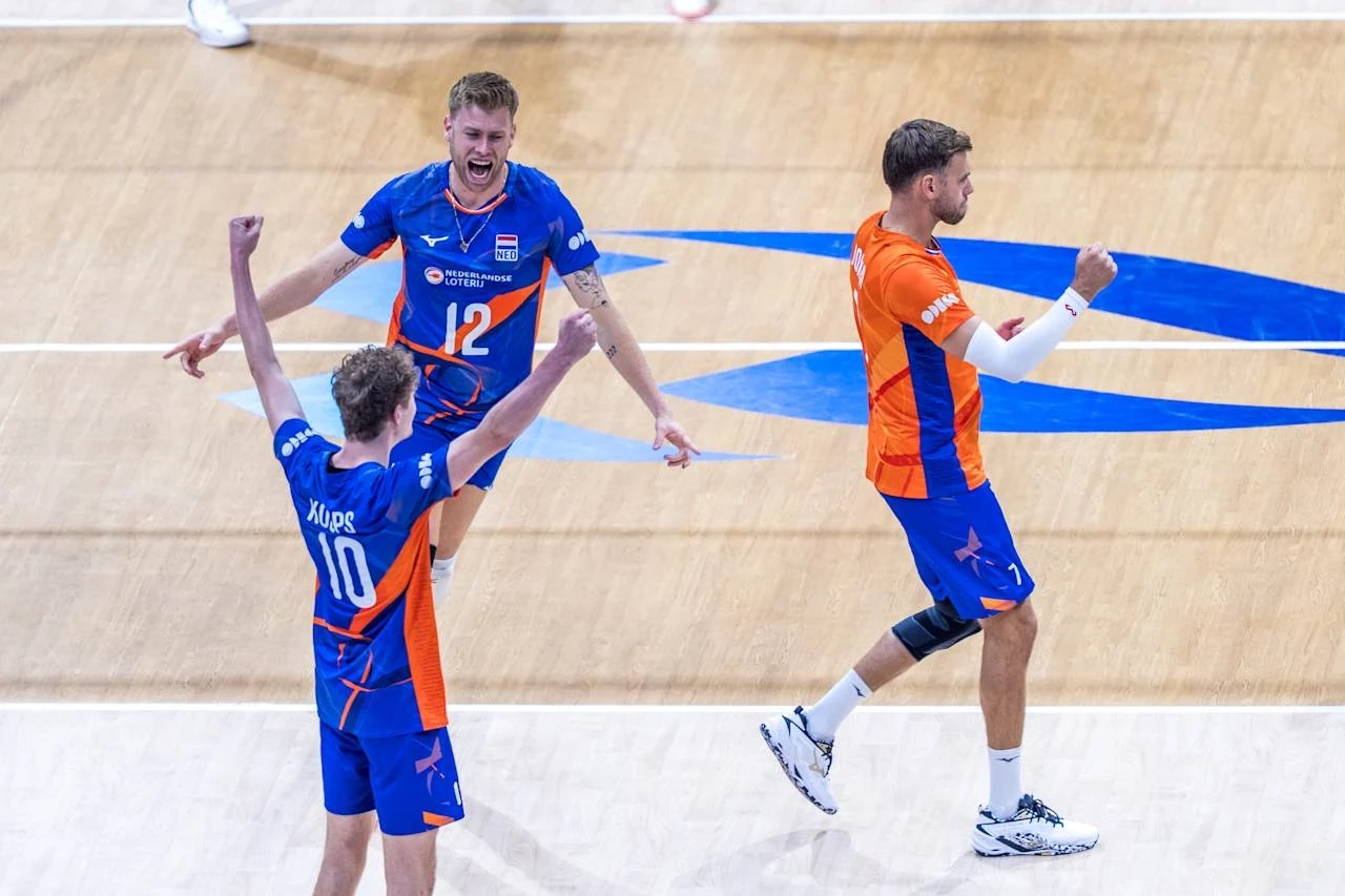 Trzech siatkarzy w pomarańczowo-niebieskich strojach świętuje zdobycie punktu na hali sportowej, uniesione ręce i ekspresja radości podkreślają emocjonujący moment meczu.