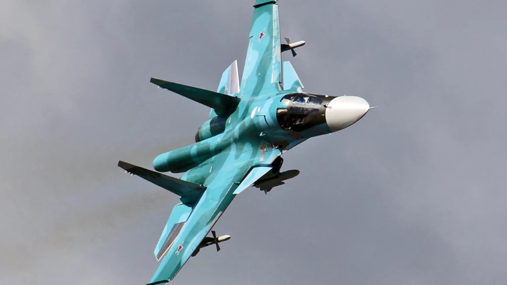 Wojskowy odrzutowiec Su-34 w locie na tle zachmurzonego nieba, maszyna ma charakterystyczną niebieską barwę oraz biały nos, widoczne szczegóły konstrukcyjne myśliwca.