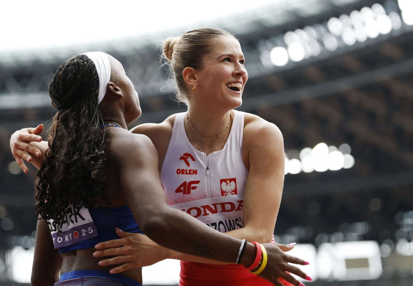 Pia Skrzyszowska< Tokio 2025 Dwie zawodniczki lekkoatletyczne obejmują się i cieszą się po ukończonym biegu, w tle widoczne są rozmyte trybuny stadionu.