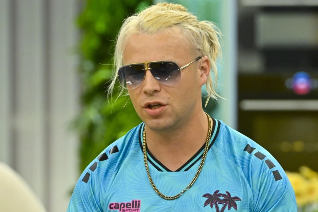 Skolim opowiedział o swojej relacji z dziećmi Mężczyzna w jasnych włosach spiętych w kucyk, ubrany w turkusową koszulkę sportową z nadrukiem palm i napisem 'capelli sport', noszący duże okulary przeciwsłoneczne i złoty łańcuch na szyi, stoi na tle zielonej roślinności oraz nowoczesnego wnętrza.