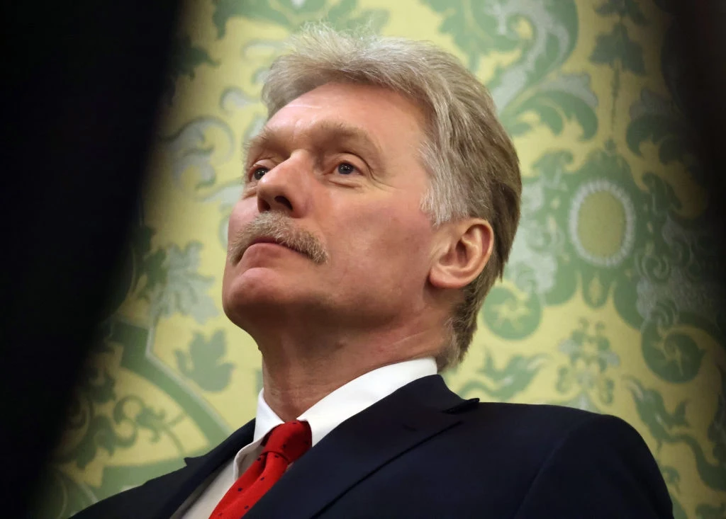 Dmitrij Pieskow: "NATO walczy z Rosją" Mężczyzna w garniturze i czerwonym krawacie, z poważnym wyrazem twarzy, blond włosy i wąsy, na tle zielono-żółtej ściany o ozdobnym, klasycznym wzorze.