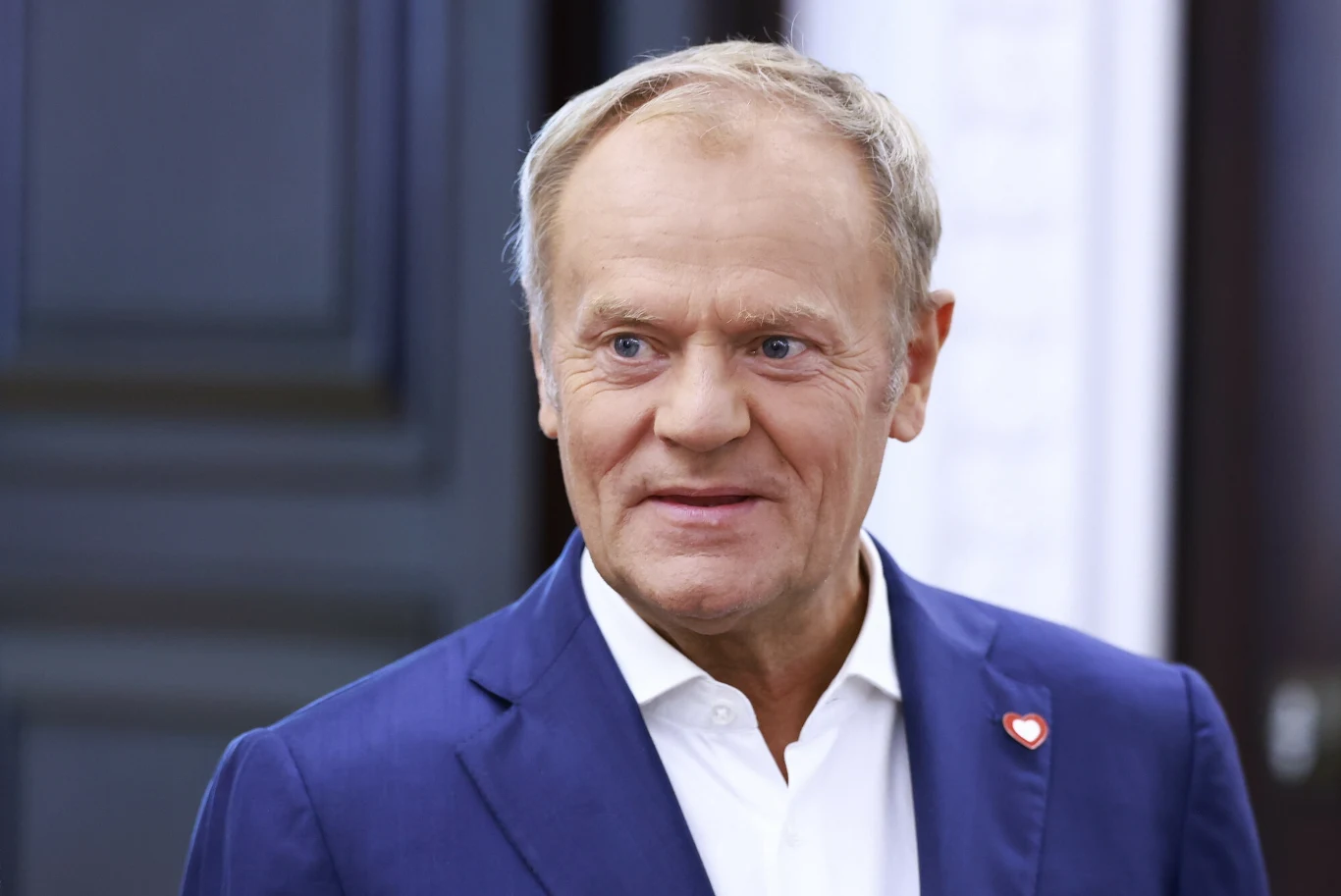 Minimalna pensja w 2026 roku ustalona. Będzie podwyżka, ale dużo niższa niż przed rokiem (zdj, ilustracyjne) Mężczyzna w średnim wieku w niebieskiej marynarce i białej koszuli z przypiętą przypinką w kształcie czerwonego serduszka, na tle ciemnych drzwi.