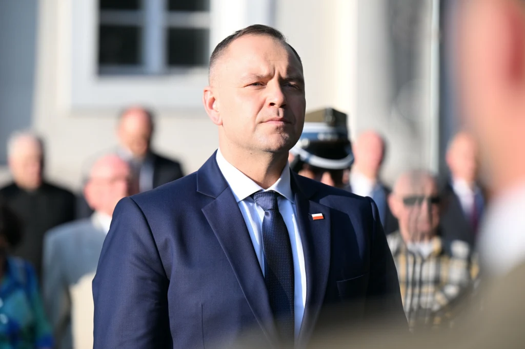 Mężczyzna w garniturze i białej koszuli z przypiętą flagą Polski stoi na tle grupy ludzi, w słonecznym świetle, z wyrazem skupienia na twarzy.
