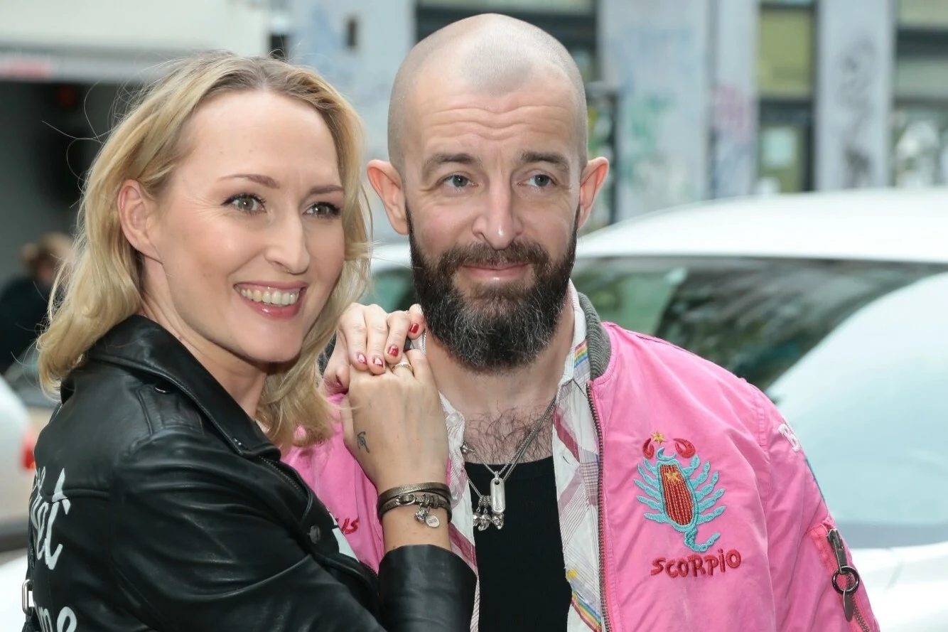 Anita Lipnicka i jej mąż Mark Gray w 2018 r. Uśmiechnięta kobieta w czarnej skórzanej kurtce i mężczyzna z brodą, ogoloną głową oraz różową kurtką z napisem 'Scorpio', trzymają się za dłonie i patrzą w stronę obiektywu, w tle rozmyte elementy miasta i biały samochód.