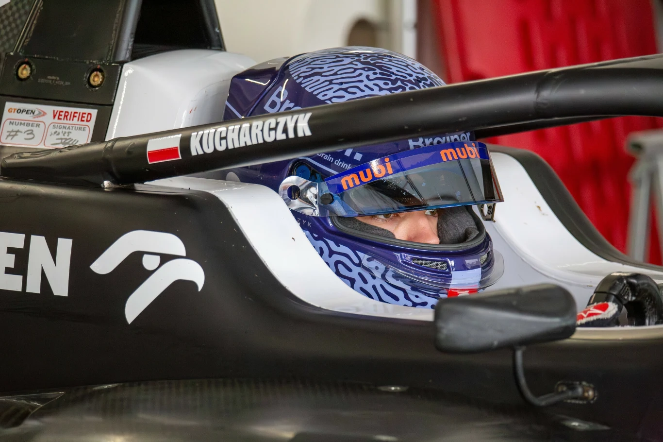 Kierowca wyścigowy w bolidzie formuły, ujęcie z bliska na jego głowę w kasku ochronnym wraz z wizjerem odbijającym światło, na aucie widoczna polska flaga i nazwisko zawodnika.