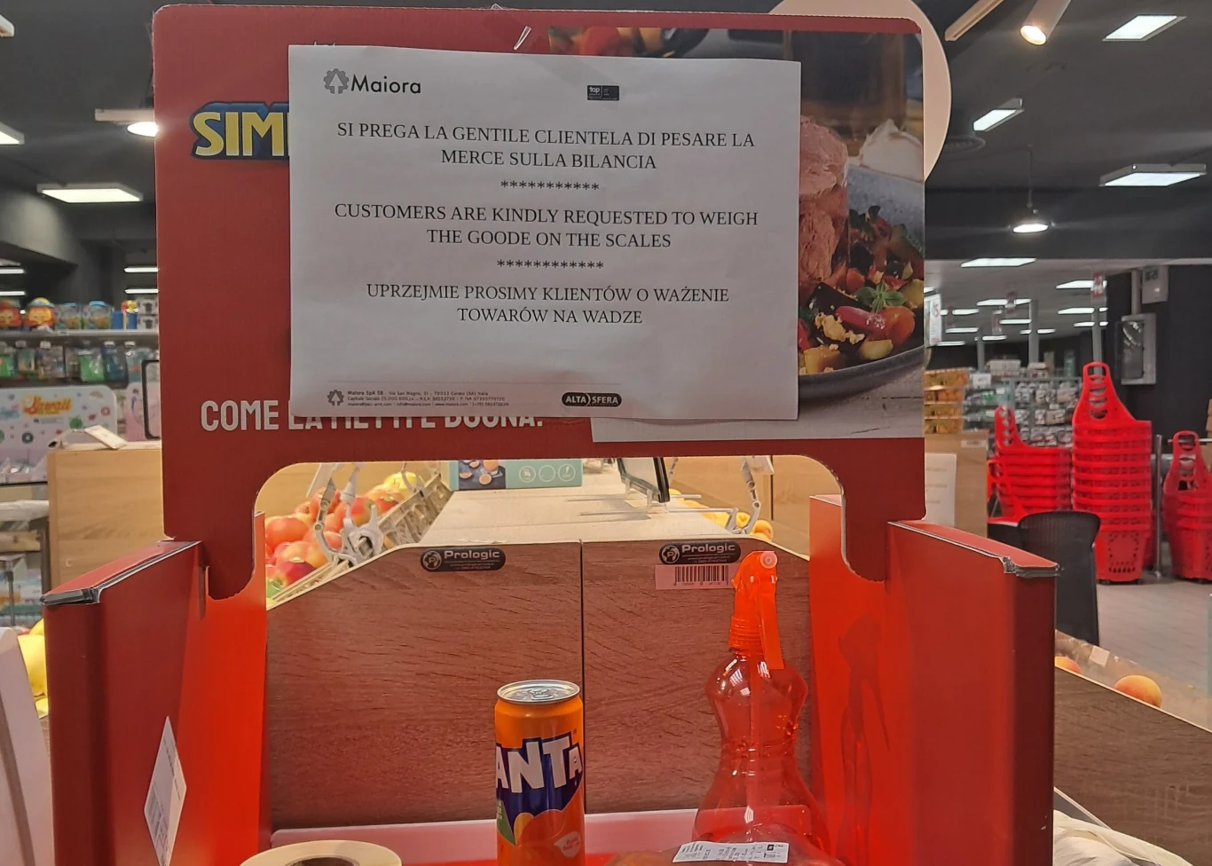 Półka sklepu spożywczego z wystawionymi napojami, na pierwszym planie puszka Fanta i butelka napoju gazowanego, nad nimi zawieszona tablica informacyjna w kilku językach z prośbą o ważenie towarów na wadze, w tle widoczne inne alejki sklepowe.
