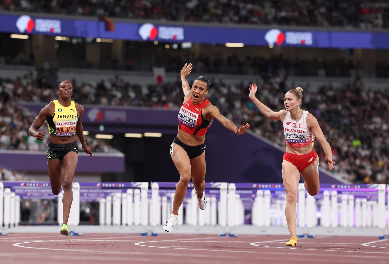 Od lewej: Danielle Williams, Ditaji Kambundji i Pia Skrzyszowska Trzy biegaczki przekraczające linię mety na stadionie lekkoatletycznym podczas rywalizacji na bieżni, widoczny tłum w tle oraz napięcie sportowe na twarzach zawodniczek.