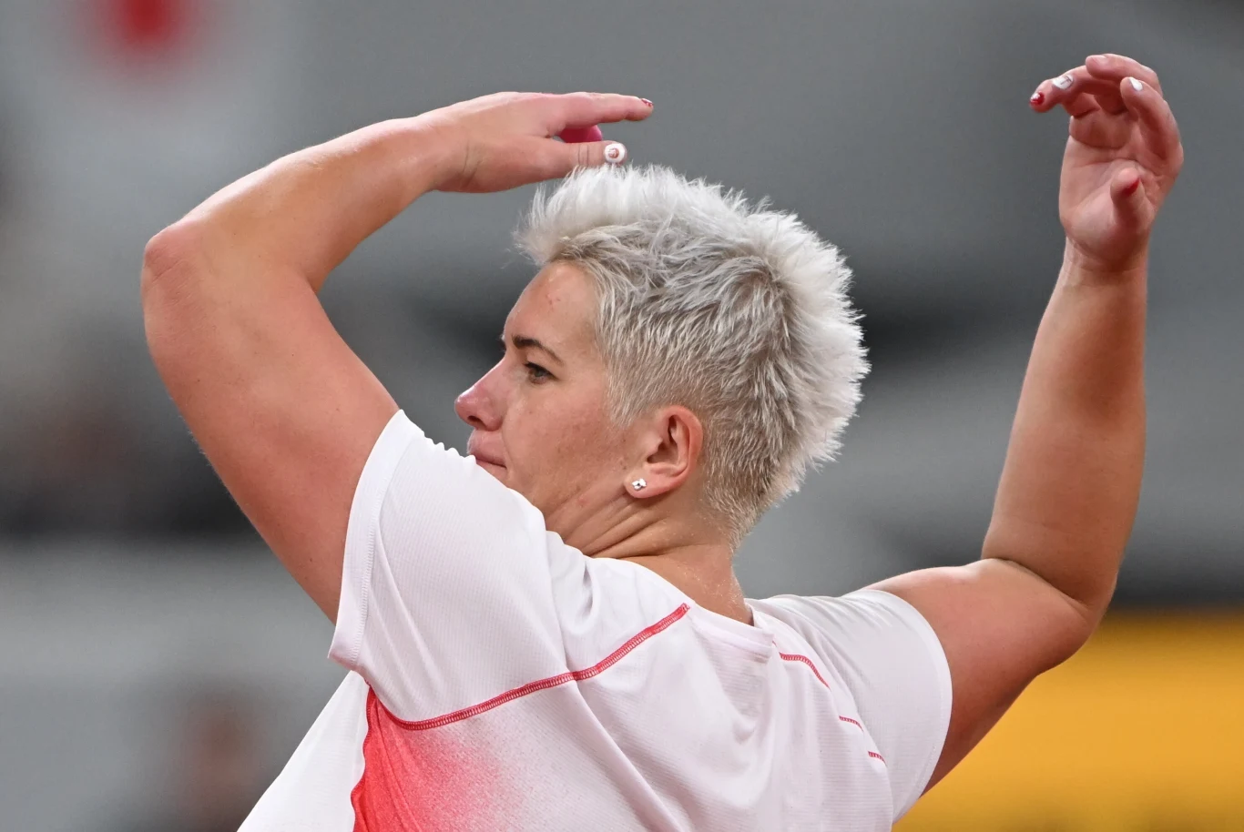 Sportowiec z krótkimi, jasnymi włosami w trakcie dynamicznego ruchu, ubrany w biało-czerwony strój, skupiony na wykonaniu zadania sportowego, sylwetka uchwycona w półobrocie.