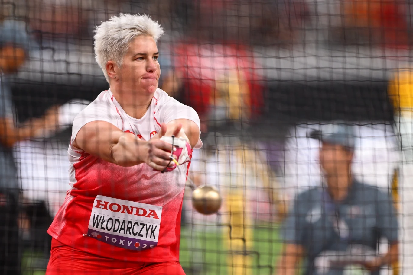 Anita Włodarczyk Zawodniczka w biało-czerwonym stroju wykonuje rzut młotem na stadionie lekkoatletycznym, w tle widoczna rozmyta postać i siatka ochronna.