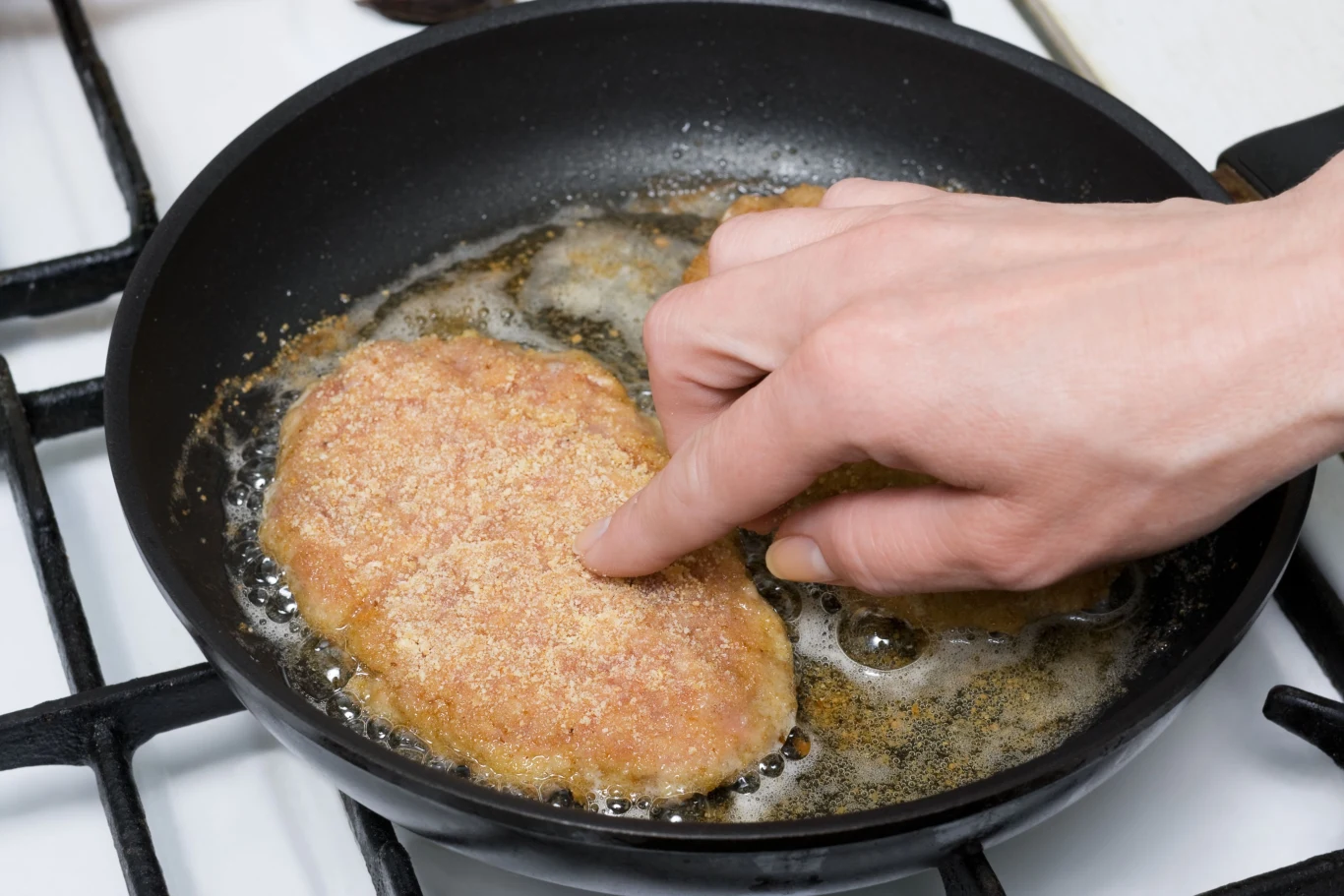 Kotlety Herkulesa możesz tradycyjnie usmażyć lub upiec w piekarniku Dłoń układa panierowany kotlet na patelni z gorącym olejem podczas smażenia.