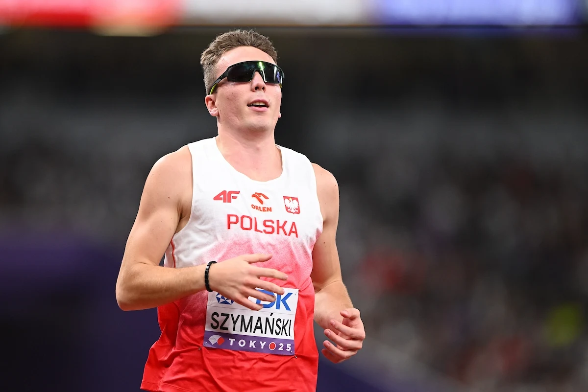 Lekkoatleta w koszulce reprezentacji Polski na tle zamazanego stadionu, biegnący w ciemnych okularach sportowych, z widocznym numerem startowym oraz nazwiskiem Szymański.