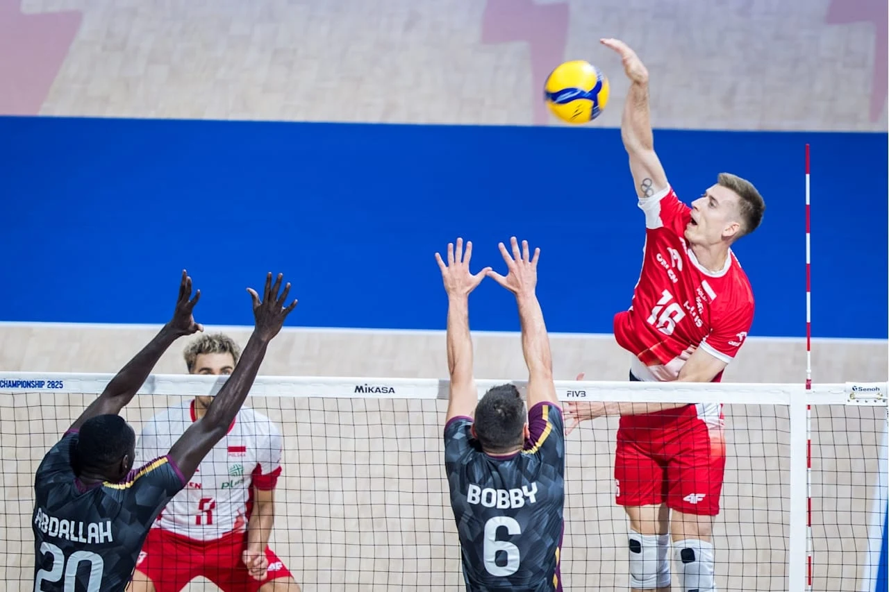 Siatkarz ubrany w czerwony strój wyskakuje i odbija piłkę nad siatką podczas meczu siatkówki, dwóch zawodników drużyny przeciwnej próbuje go zablokować podnosząc ręce.