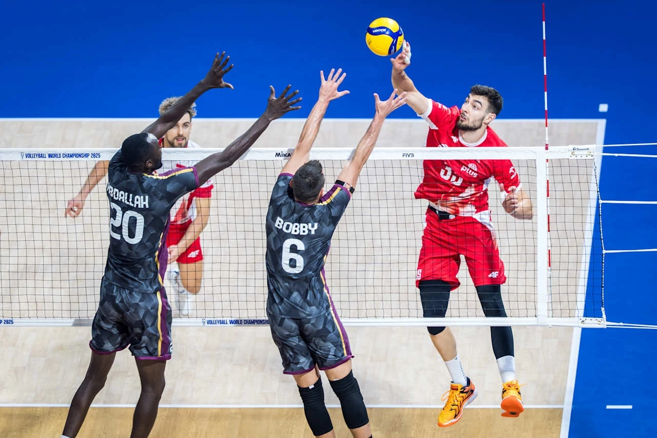 Zawodnik w czerwonym stroju wykonuje atak podczas meczu siatkówki halowej, a dwóch zawodników w ciemnych strojach z numerami 20 i 6 próbuje zablokować piłkę przy siatce. Piłka znajduje się nad siatką w trakcie akcji.