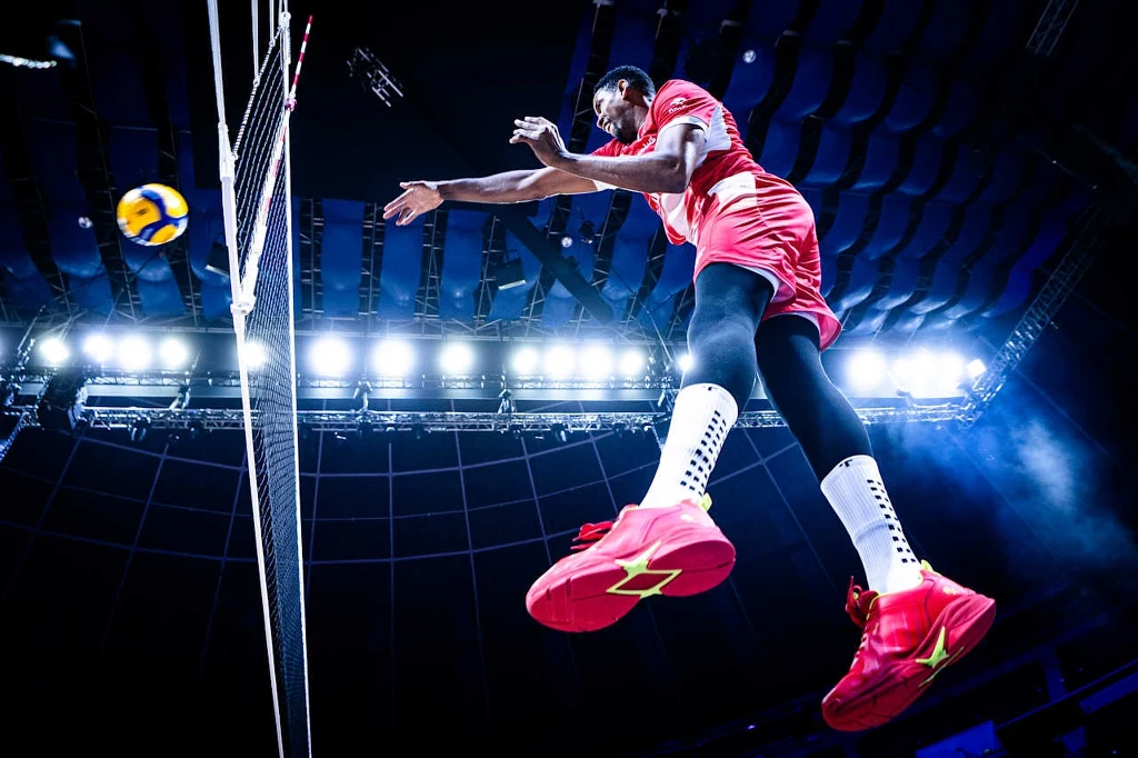 Wilfredo Leon Zawodnik siatkówki w dynamicznym skoku nad siatką, przygotowuje się do wykonania ataku, a w tle jasne światła hali sportowej podkreślają energię momentu.