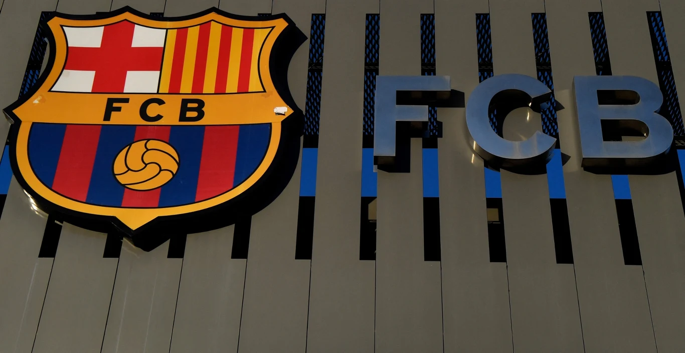 Herb klubu piłkarskiego FC Barcelona oraz duże, metaliczne litery FCB na jasnym tle fasady budynku.