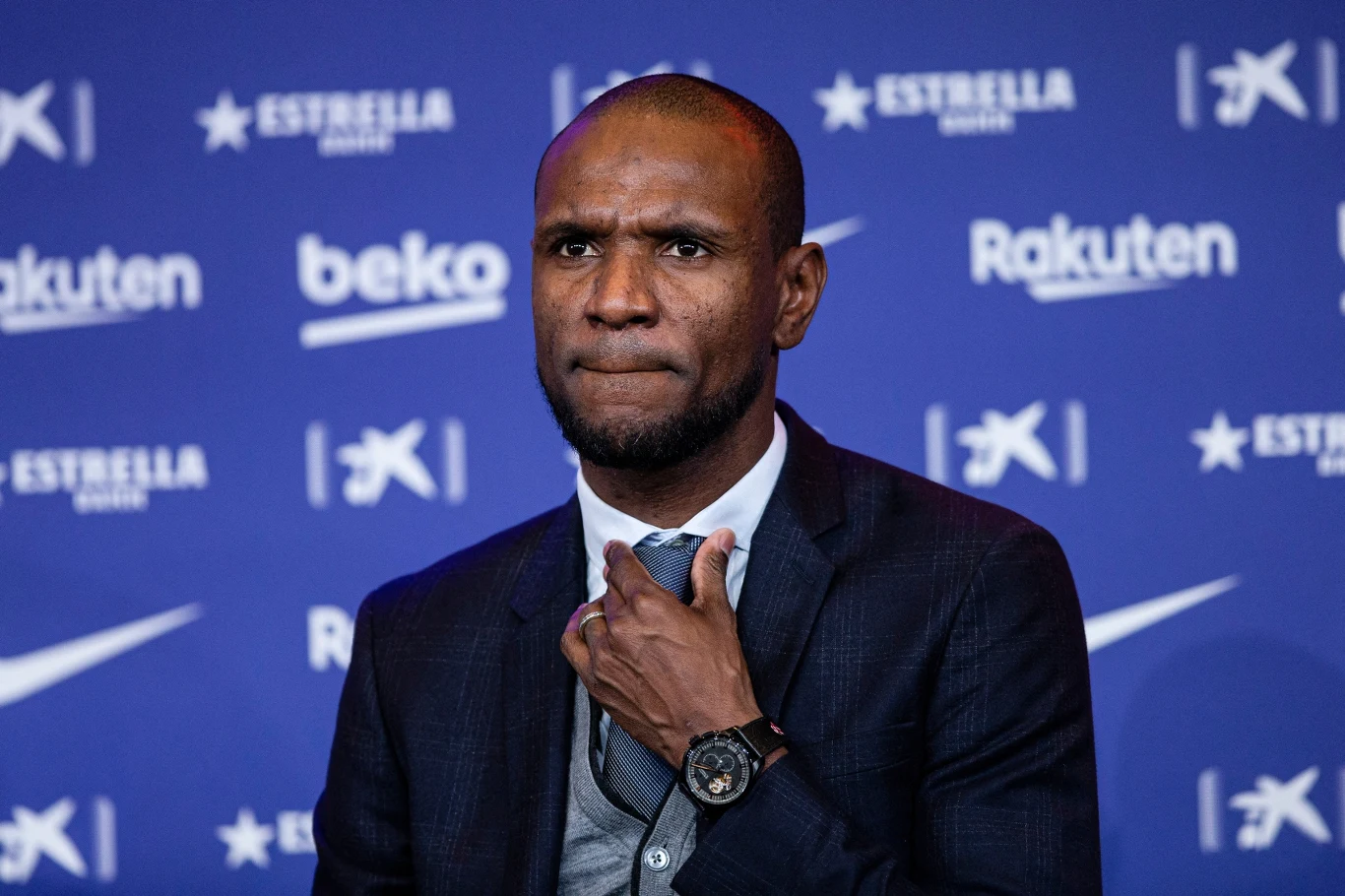 Eric Abidal Mężczyzna w garniturze stojący na tle niebieskiej ścianki z logotypami firm i sponsorów, poprawiający krawat, z poważnym wyrazem twarzy.