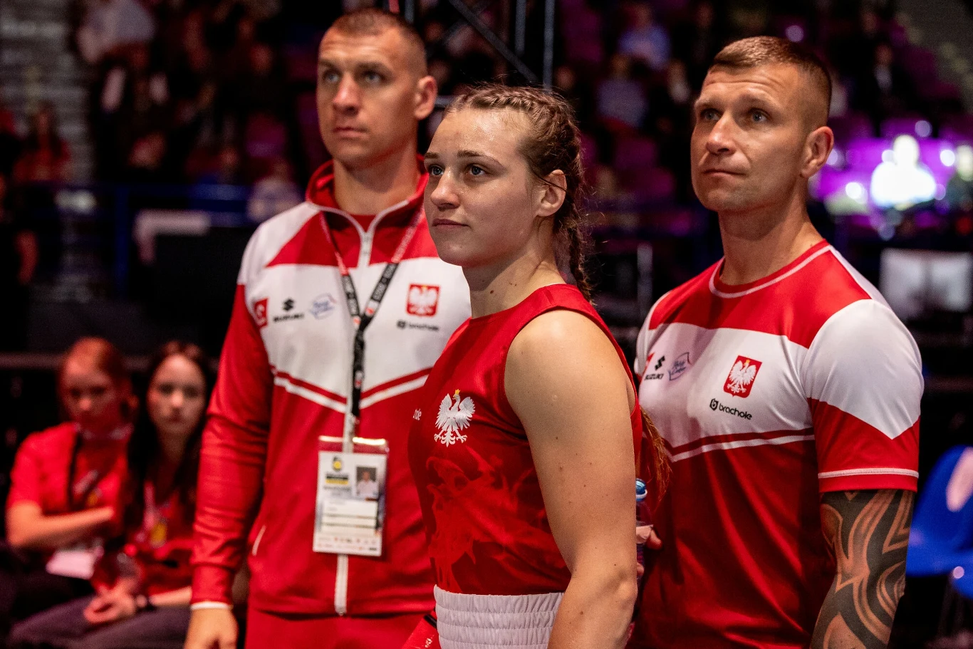 Julia Szeremeta z trenerami Trójka sportowców w czerwono-białych strojach z orzełkami na piersi stoi wśród ludzi, dwie osoby w sportowej odzieży reprezentują Polskę, kobieta na pierwszym planie w stroju bokserskim przygotowuje się do zawodów, atmosfera skupienia i oczekiwania.