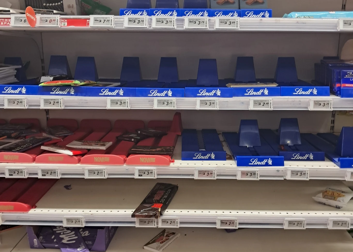 Puste półki sklepowe z niewielką liczbą pozostałych tabliczek czekolady, w większości marki Lindt i niektórych innych, z widocznymi cenami na etykietach, świadczące o dużym popycie lub niedoborze produktowym.